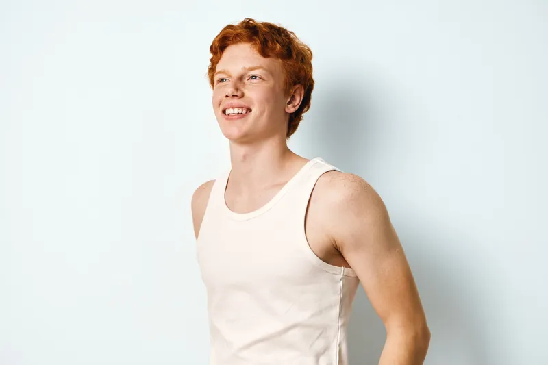 Young man with red hair wearing white tank top standing against white background smiling and looking away. Casual confident male portrait with relaxed expression and bright lighting.