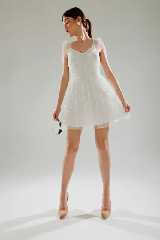 Young woman in a white elegant dress poses confidently holding sunglasses. Studio shot on a neutral background. Concept of woman beauty fashion, style, and elegance.