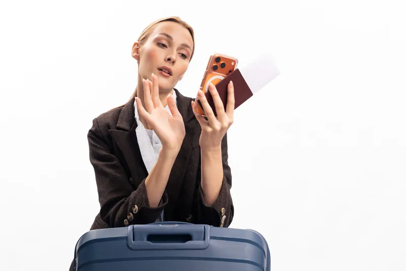 Young woman holding passport and smartphone waves hand while standing behind suitcase. Travel concept for vacation, airport, and journey planning on white background.