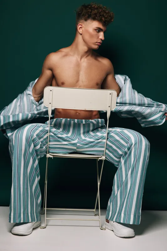 Young man sitting on white metal chair with striped pants and open shirt removing jacket. Model with curly hair looks sideways in studio against green background with shadow on floor.