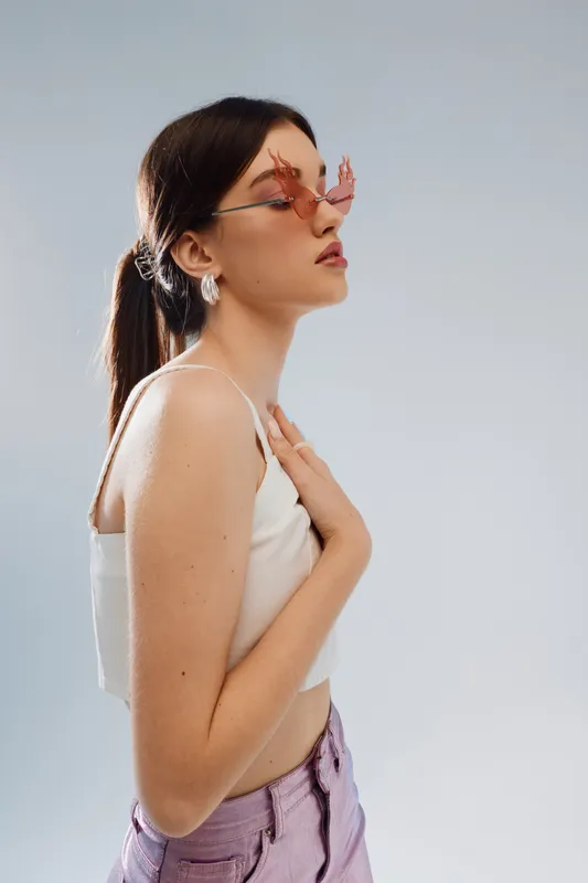 Young woman in white top and purple pants wearing pink sunglasses poses in profile against a light background. Concept of beauty, fashion, and style.
