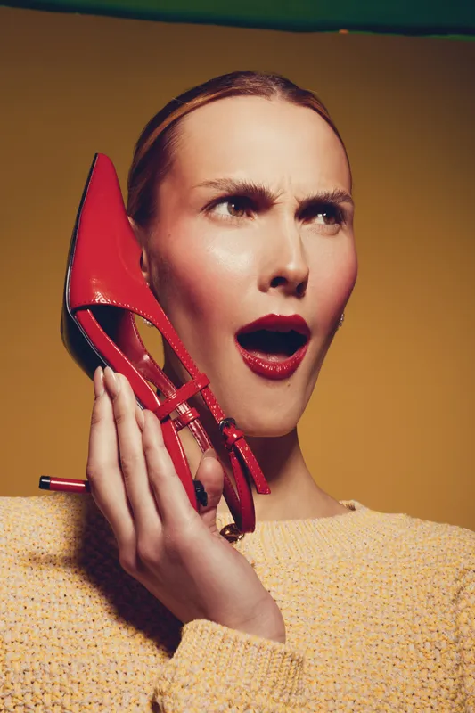 Surprised woman with red lipstick holds red high heel shoe near her ear, wearing yellow sweater. Studio portrait on yellow background with expressive facial expression and fashion concept.