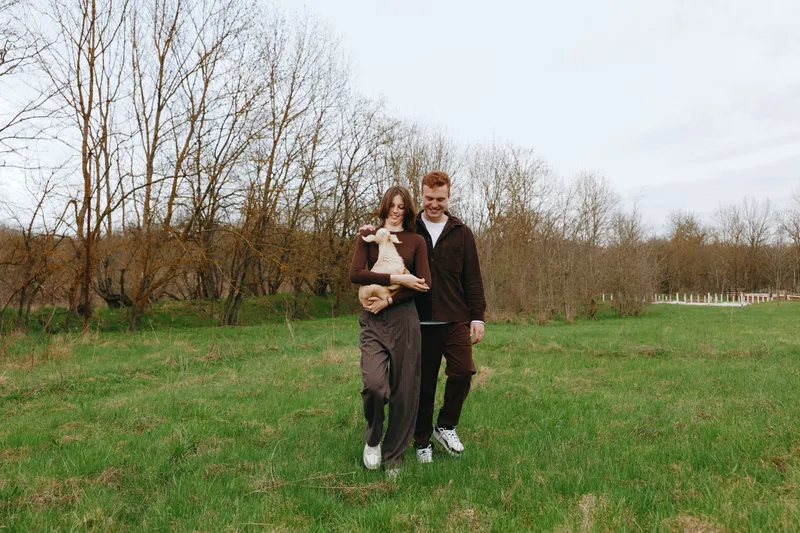 Casual couple strolls through an open field with a playful goat kid, enjoying a relaxed lifestyle moment, natural light, candid mood, rustic scenery, calm atmosphere