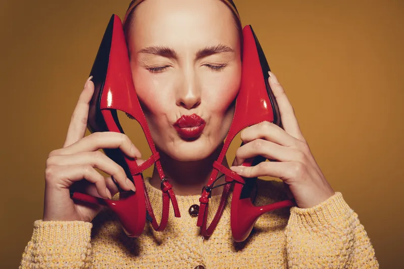 Young woman with closed eyes holds red high heels near face, wearing yellow sweater. Concept of fashion, style, beauty, and playful mood in studio with warm background.