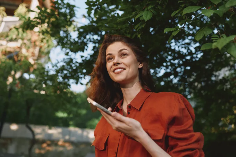 Young woman in a red shirt smiles while holding a phone outdoors, enjoying a sunlit moment in a park setting among lush greenery, her relaxed posture and joyful gaze reflecting everyday life