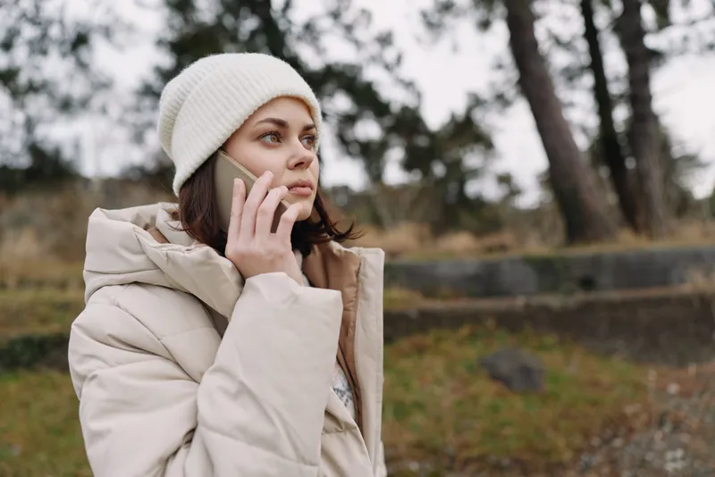 Woman, phone, outdoor, cold, winter, coat, jacket, talking. A woman in a beige puffer coat and knit hat speaks on a mobile phone in a chilly park setting with bare trees and overcast sky, conveying
