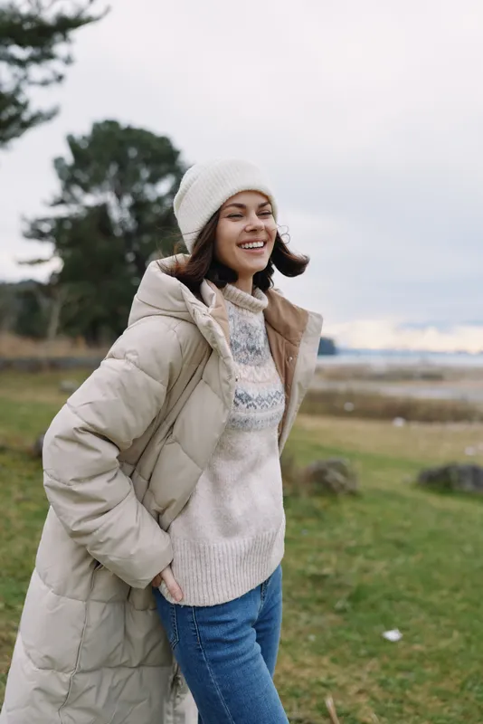 Woman in a long winter coat and knit sweater smiles outdoors in a grassy field with trees and cloudy sky, conveying cozy seasonal style and relaxed mood.