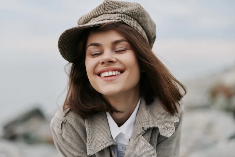 Smiling young woman with closed eyes wears beige cap and coat outdoors. She looks happy and relaxed in natural light with blurred background. Concept of joy, fashion, and freedom.
