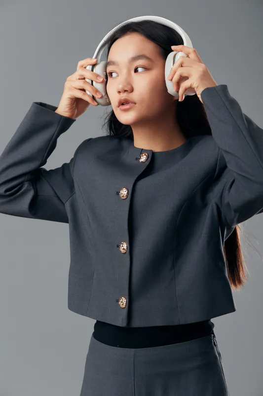 Young woman wearing gray jacket and wireless headphones indoors looking aside with thoughtful expression. Casual style, long black hair and modern technology for music or communication.