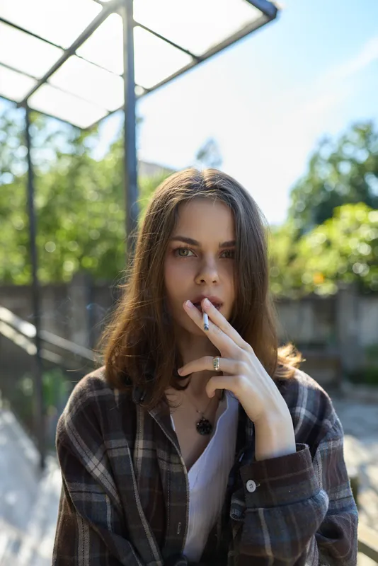 Young woman outdoors wearing a plaid shirt, cigarette in hand. Casual lifestyle portrait on a sunny day with soft natural light and a relaxed mood in frame