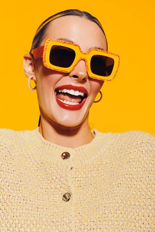 Smiling woman wearing large orange sunglasses and yellow sweater poses against bright yellow background. Concept of fashion, summer style, happiness, and vibrant colors.