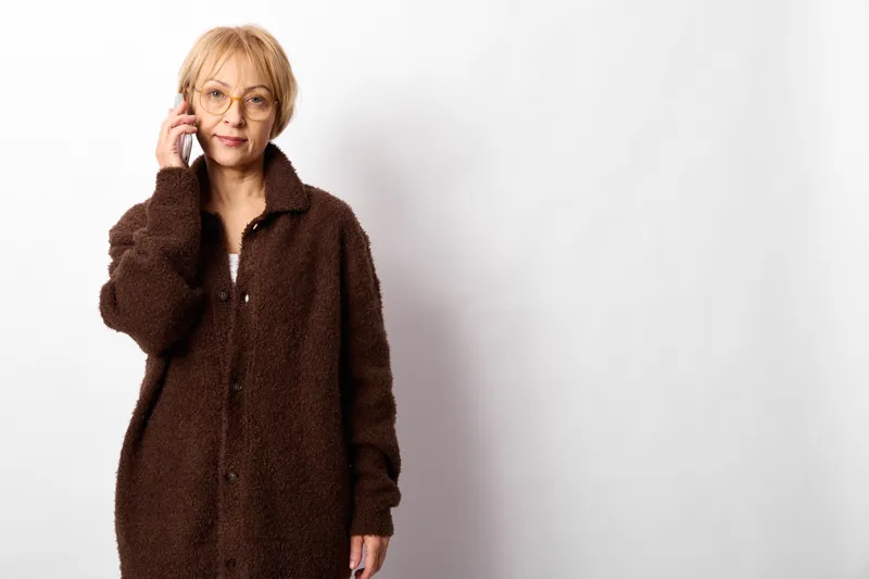 Woman with glasses, senior in brown coat, holding phone to ear, studio portrait indoors on white background, soft light, calm communication