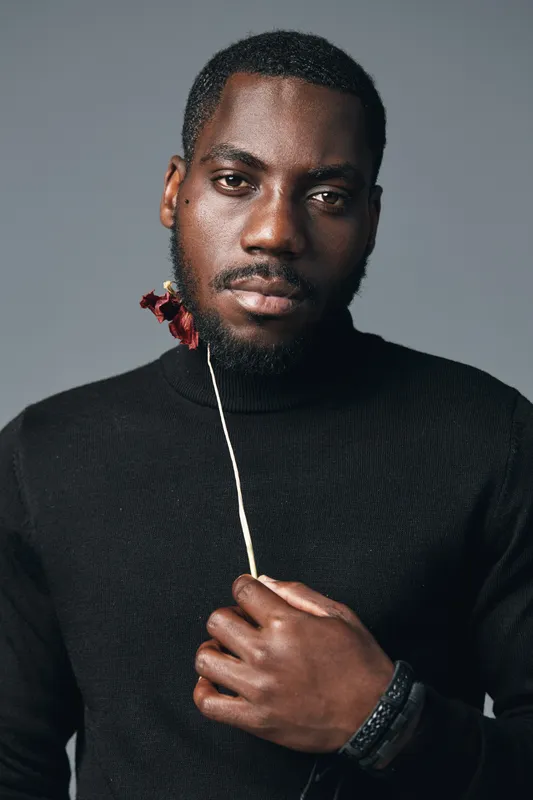 African man with dark skin holding red flower in creative conceptual visual style, portrait with serious expression wearing black sweater on gray background.
