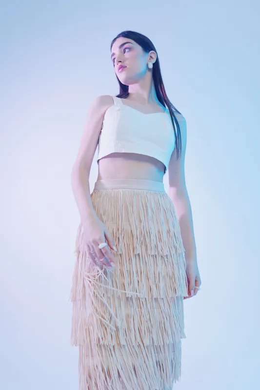 Young woman in a white crop top and beige fringed skirt poses against a light background. Soft lighting highlights her beauty and fashion style. Concept for modern fashion and elegance.