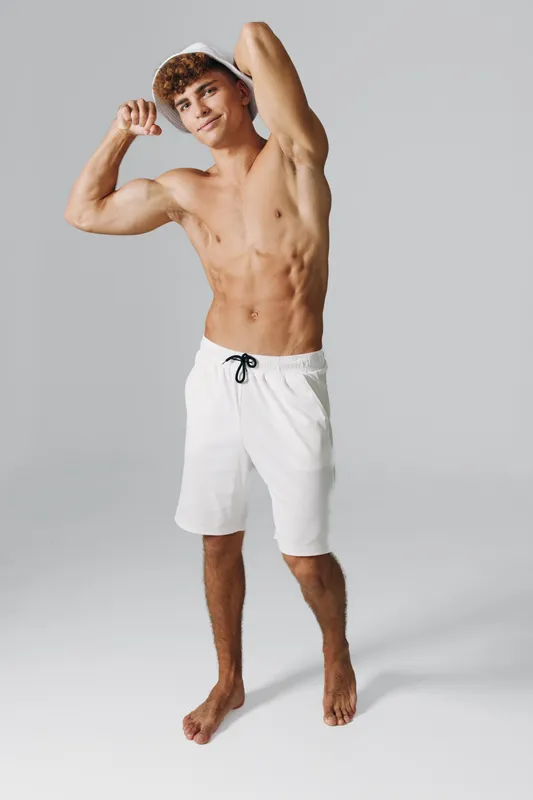 Young man posing shirtless in white shorts and bucket hat showing muscular body with flexed biceps on gray studio background. Adult guy standing barefoot, confident and smiling.