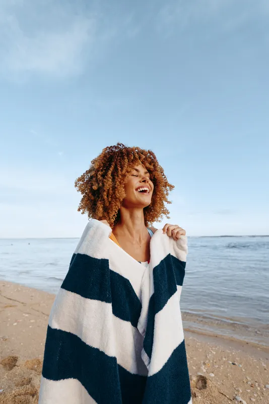 Woman smiling curly hair on beach by ocean under sky, wrapped in striped blanket, outdoors in soft daylight, relaxed posture, sea breeze, gentle waves, natural colors, joyful laughter