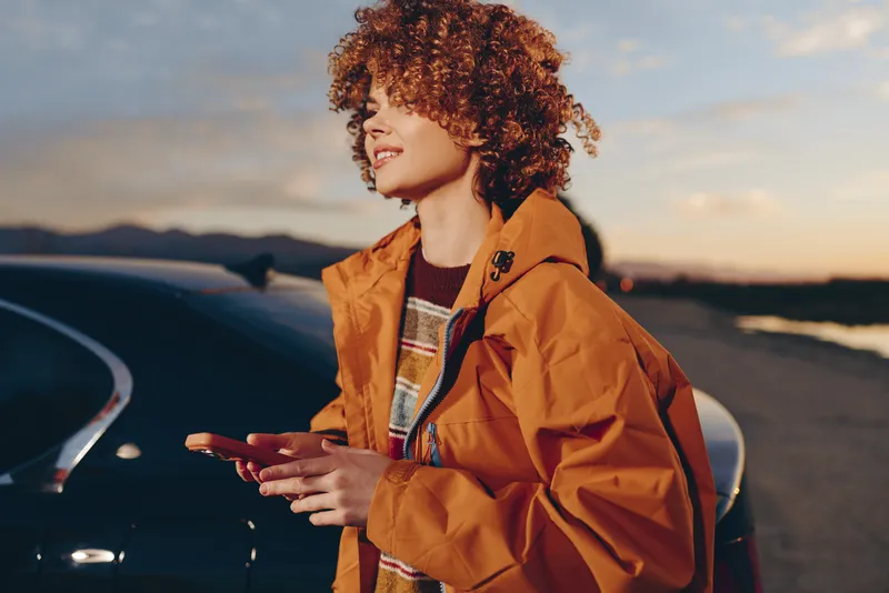 Woman lifestyle portrait outdoors at sunset, curly hair, orange jacket, standing by car, relaxed smile, hands near pockets, travel mood, warm evening light, roadside view, golden hour sky