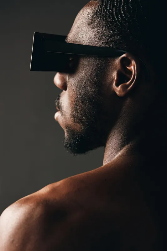 African man with dark skin wearing futuristic black glasses in creative conceptual visual style. Closeup portrait with dramatic lighting and artistic shadow on bare shoulder.