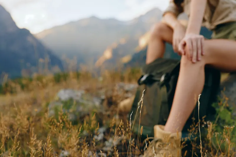 Woman lifestyle traveler resting on rock in nature with mountains background wearing boots and shorts enjoying outdoor adventure and peaceful moment in wilderness.