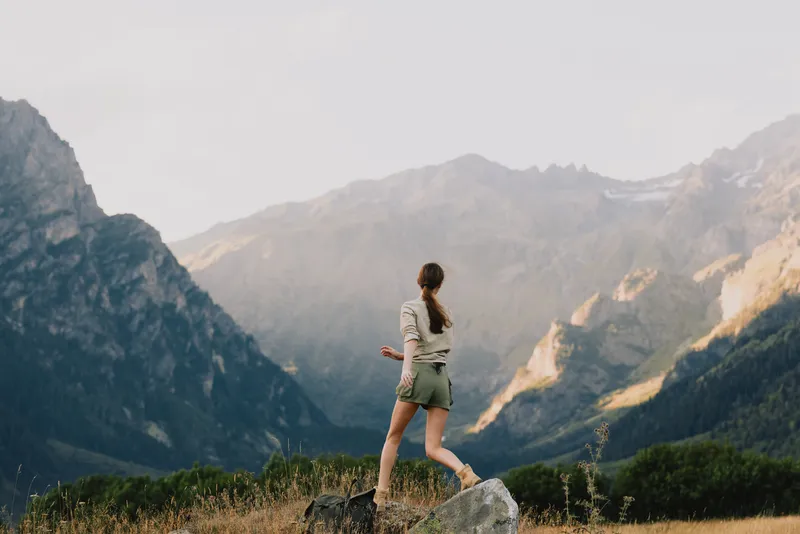 A woman stands on a rocky trail overlooking rugged mountains, enjoying outdoor life and fresh air. The scene conveys adventure, balance, resilience, and appreciation for nature.