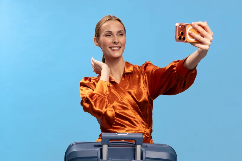 Young woman in orange satin blouse takes selfie with smartphone while standing behind blue suitcase. Studio portrait on blue background. Concept of travel, tourism, and modern lifestyle.