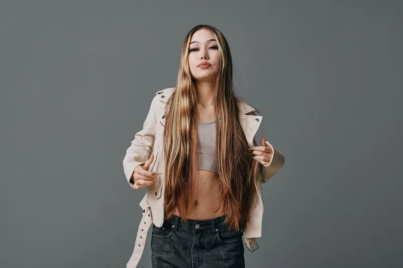 Young woman with long braided hair wearing casual beige jacket and crop top posing confidently against gray background in studio session for fashion and style photography.