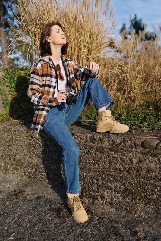 Woman outdoors with camera resting on knee, wearing plaid shirt, jeans and boots, sitting on a stone ledge among tall grasses as sunlight highlights a relaxed moment.