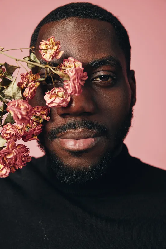 Creative conceptual portrait of African man with dried pink flowers covering one eye against pink background. Visual style emphasizes contrast, texture, and artistic expression.