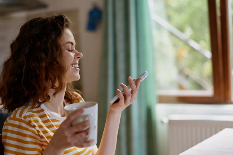 Smiling young woman with curly hair talks on her phone while holding a mug in a bright sunlit room by a window
