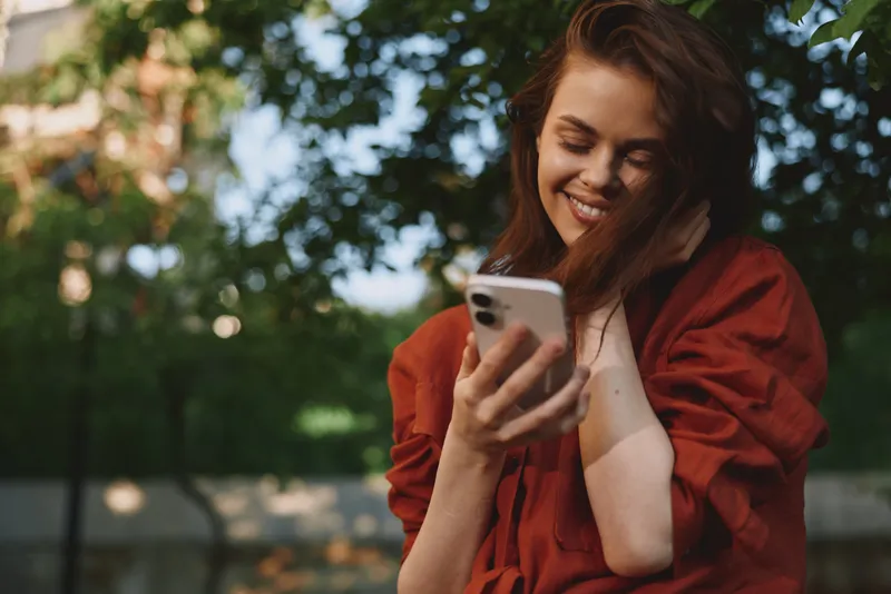 Young woman in a red blouse stands outdoors, smiling at her phone as sun dappled leaves frame a candid moment of everyday connection and relaxed lifestyle