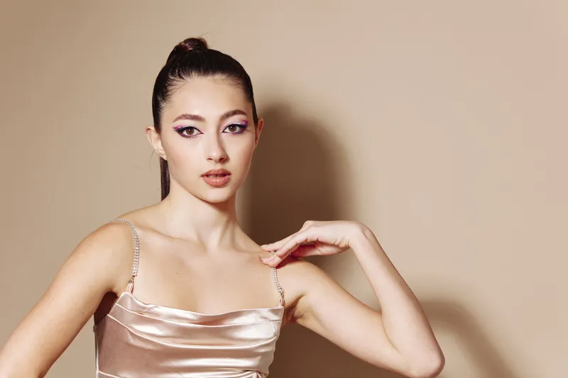 Young woman in a satin dress poses with elegant makeup and sleek hairstyle on beige background. Beauty fashion concept emphasizing creative style and modern glamour.