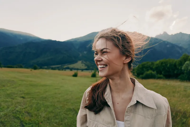 Young woman with long hair smiles joyfully while standing in a green meadow with mountains in the background during a bright day.