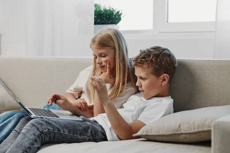 Two children sitting on a sofa using a laptop together, focused and engaged in a bright living room. Concept of childhood, technology, and learning at home.