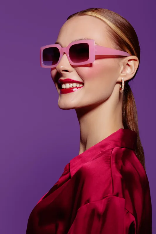 Smiling woman with pink sunglasses and red lipstick wears red satin blouse against purple background. Fashion portrait with bright makeup and trendy accessories in studio lighting.