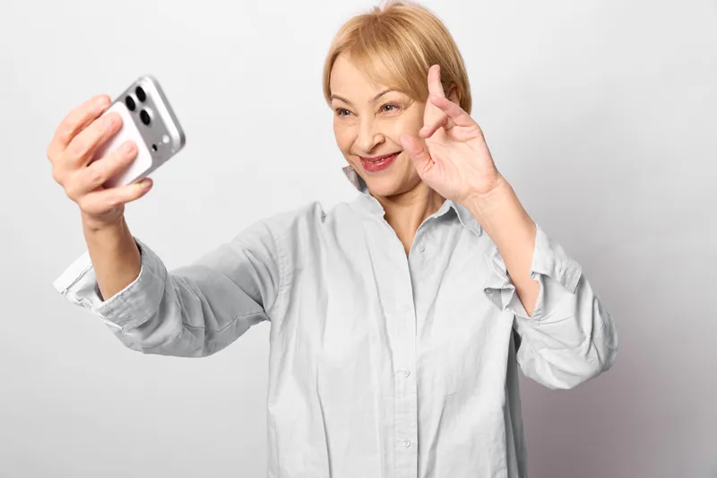 Senior woman smiling indoors, holding smartphone for selfie, making peace gesture with fingers, bright studio light, calm relaxed mood, modern technology