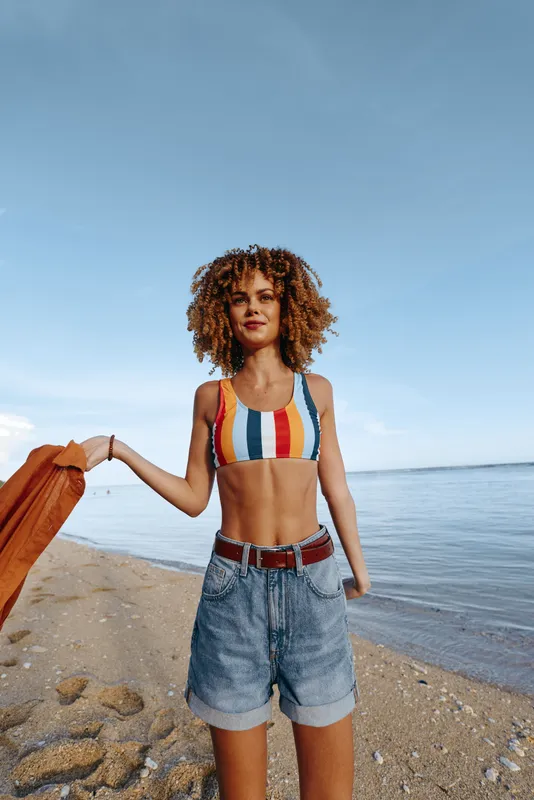 Woman on a sunny beach wearing a striped bikini top and denim shorts, confident.