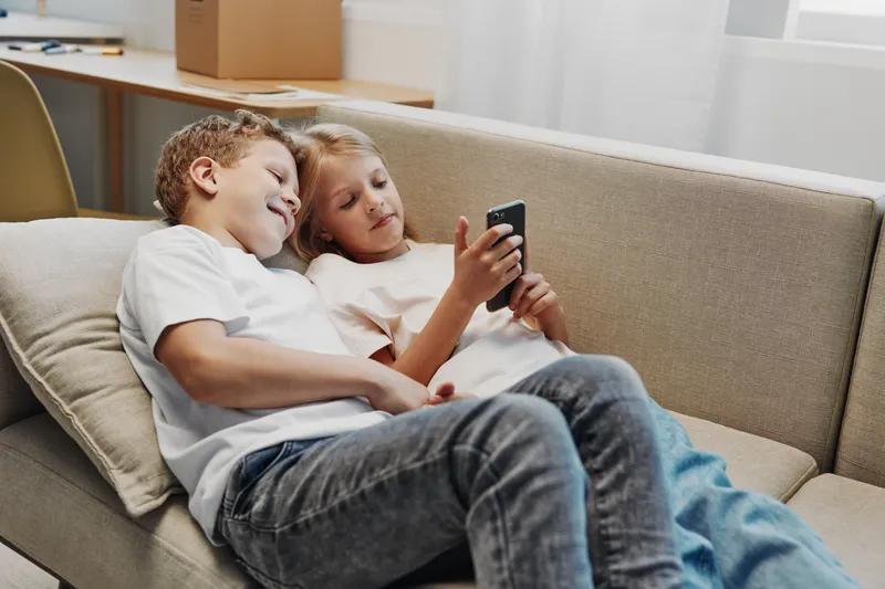 Two children relax on a couch, sharing a smartphone and smiling together in a bright living room. Concept of friendship, technology, leisure, and childhood bonding.