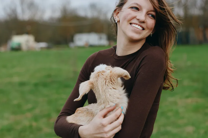 Cheerful woman outdoors, smiling while holding a goat kid in a sunlit meadow, casual sweater, relaxed posture, natural colors, warm breeze, pastoral lifestyle, gentle connection with animals