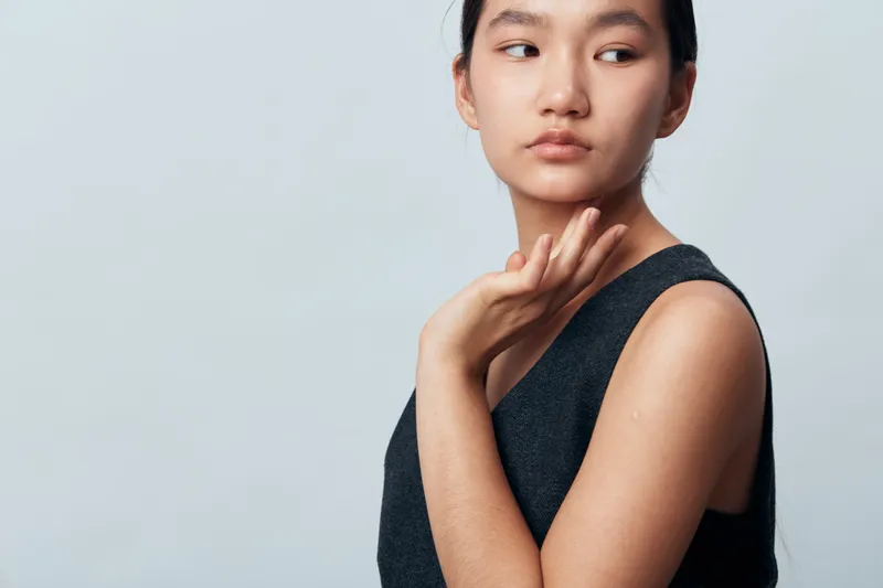 Young Asian woman with clear skin wearing dark sleeveless top posing on grey background with serious expression and hand raised near neck in beauty portrait concept.
