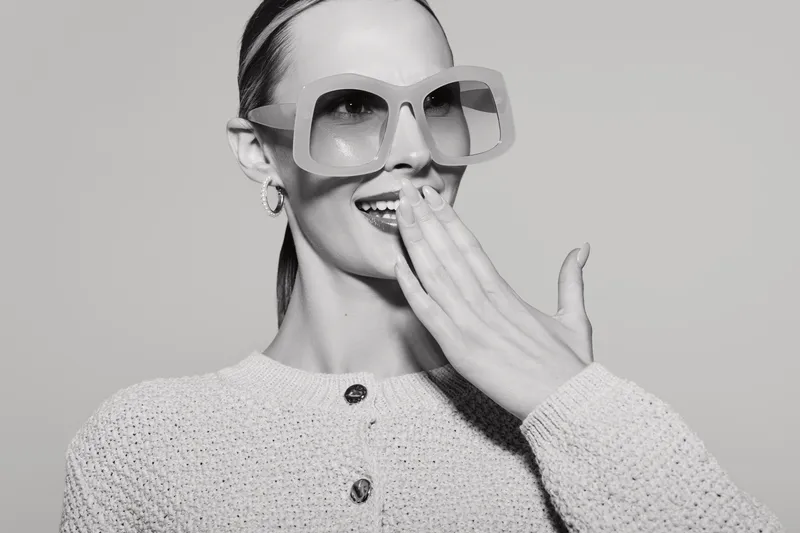A smiling caucasian woman with pale skin wearing oversized sunglasses and a knitted sweater covers her mouth with her hand. Studio portrait on grey background with soft lighting.