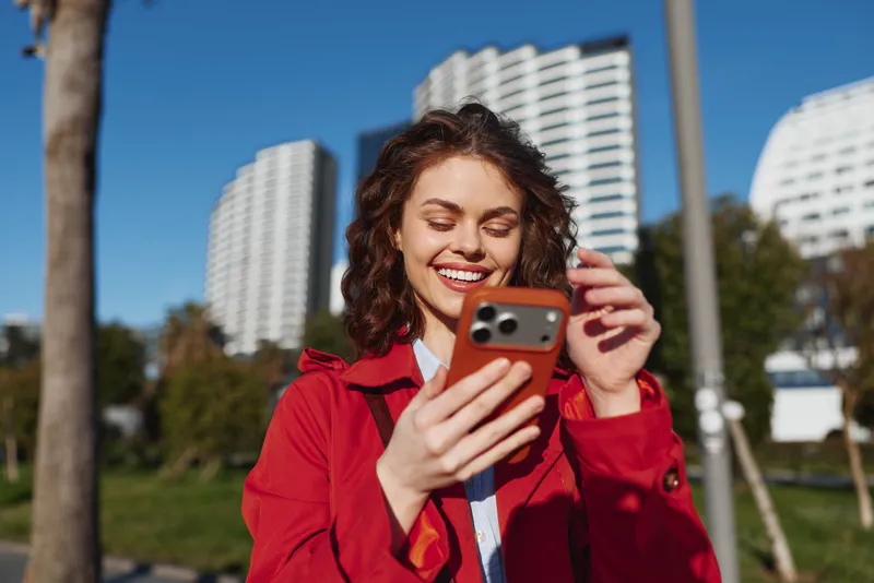 Young woman in red coat smiling while using smartphone outdoors in city park with modern buildings in background on sunny day