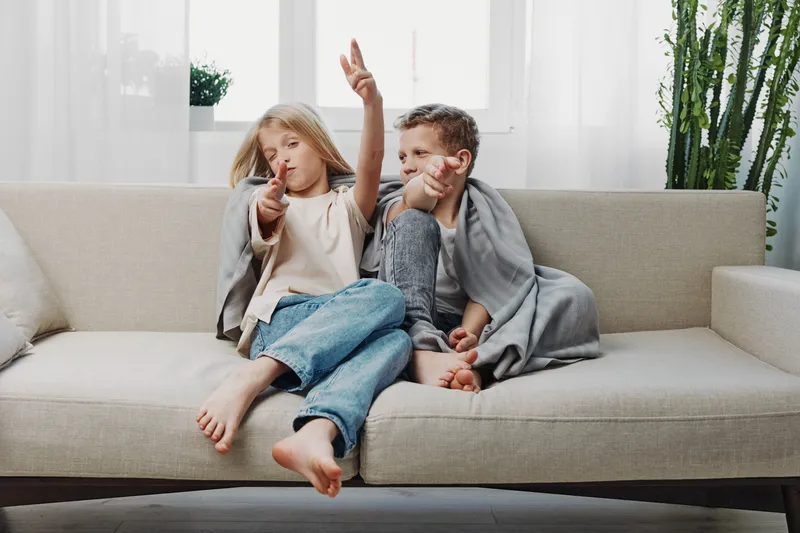 Two playful children sitting on beige sofa covered with gray blanket, pointing fingers like guns, enjoying relaxed moment in bright living room with plants. Concept of childhood fun.