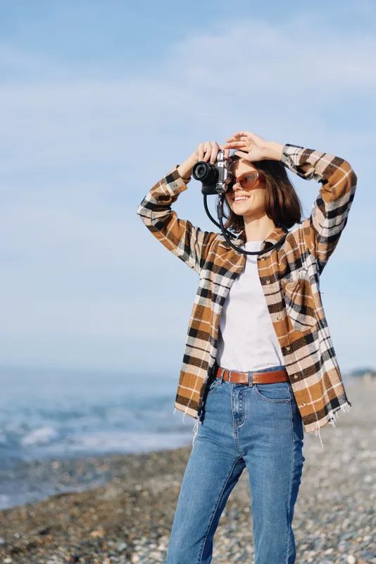 Photographer, woman with camera capturing coastal scenery under clear sky, wearing plaid shirt and jeans, standing on a rocky beach with sea breeze and distant horizon