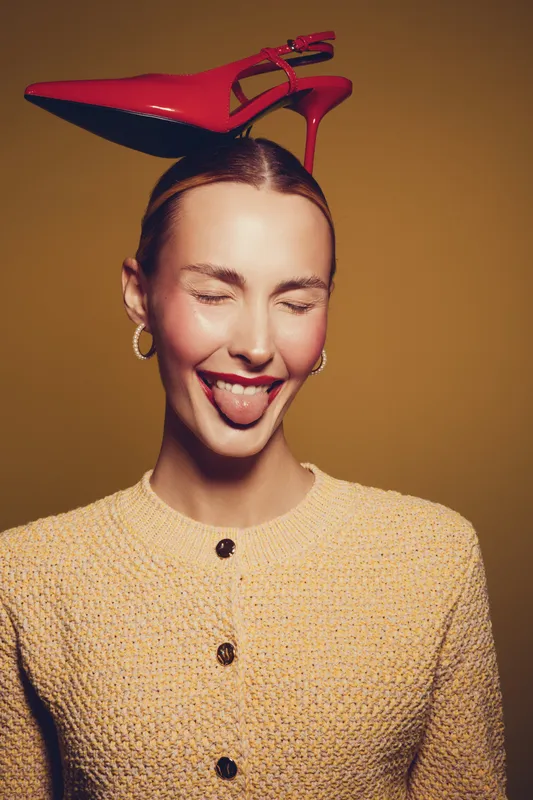 Young woman with closed eyes and red lipstick sticks out tongue while balancing red high heel shoe on head. She wears yellow textured jacket and gold hoop earrings on brown background.