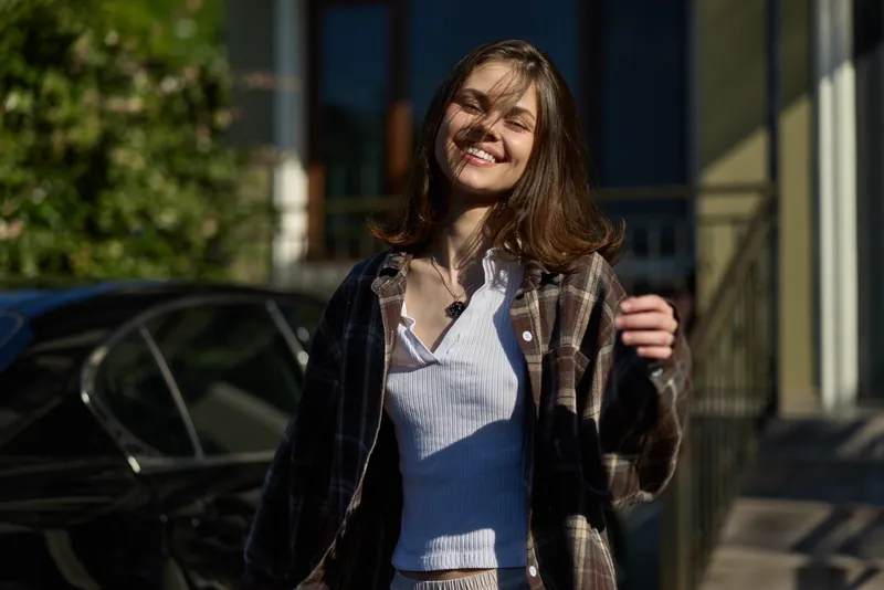 Bright casual portrait of a smiling woman enjoying outdoor lifestyle, wearing a plaid jacket over a striped shirt, natural daylight, urban setting, relaxed candid moment