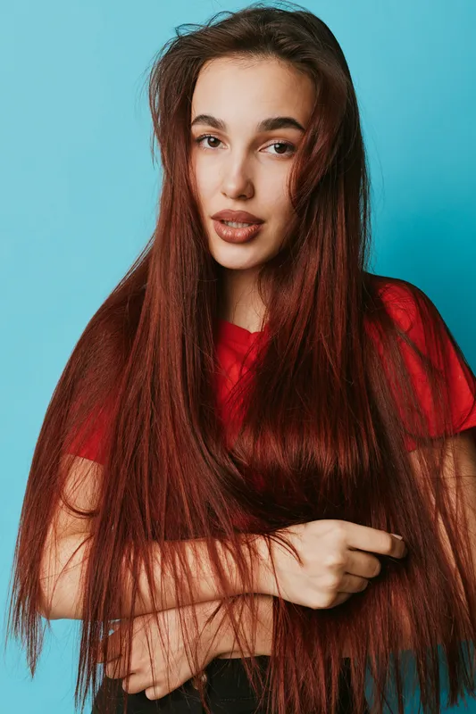 Young woman with long straight brown hair wearing red shirt posing against blue background, looking at camera with natural expression and relaxed posture in studio portrait.