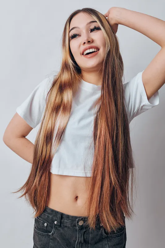 Young woman with long straight hair wearing white cropped t shirt and jeans, posing with hand behind head and smiling in studio with plain background.