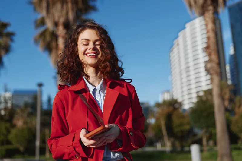 Smiling young woman with curly hair wearing a red coat and white shirt stands outdoors in a sunny park setting with modern buildings in the background.