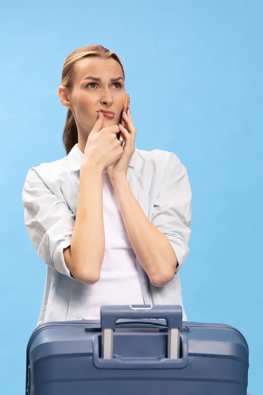 Thoughtful woman in casual white shirt holds smartphone to ear, looks up with hand on chin. Blue suitcase in foreground on blue background. Concept of travel planning and decision making.