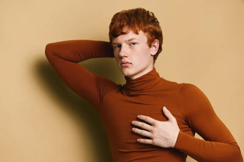 Young man with red hair wearing brown turtleneck posing with hand on chest and another behind head against beige background for fashion portrait studio.